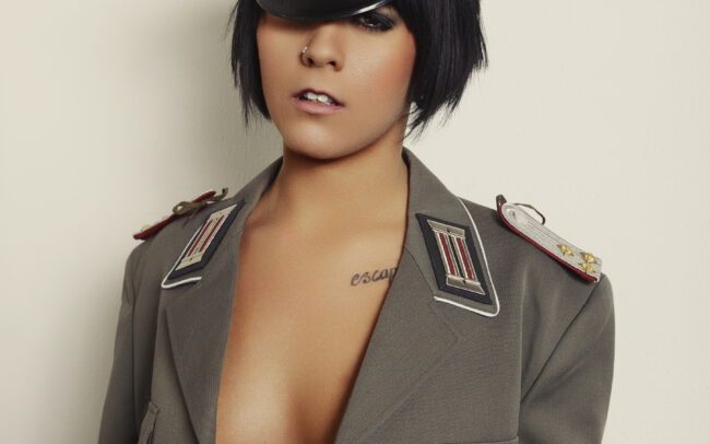 Fine art studio portrait of a woman in a DDR officer’s jacket and peaked cap, close framing — by Menno van der Meulen