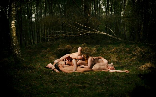 Fine art photograph by Menno van der Meulen, “Fields of Joy” — a group of nude figures embracing in a mossy forest clearing, warm skin tones framed by deep green woodland.