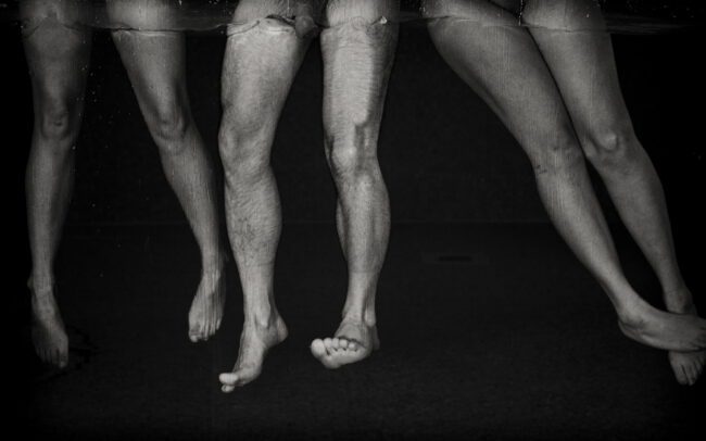 Fine art black-and-white photograph of submerged legs hovering beneath the water’s surface, by Menno van der Meulen