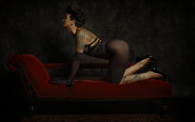 Fine art photograph of Miss V kneeling on a red velvet chaise longue, tattooed, in a sheer black dress and heels, dramatic low-key lighting — by Menno van der Meulen.
