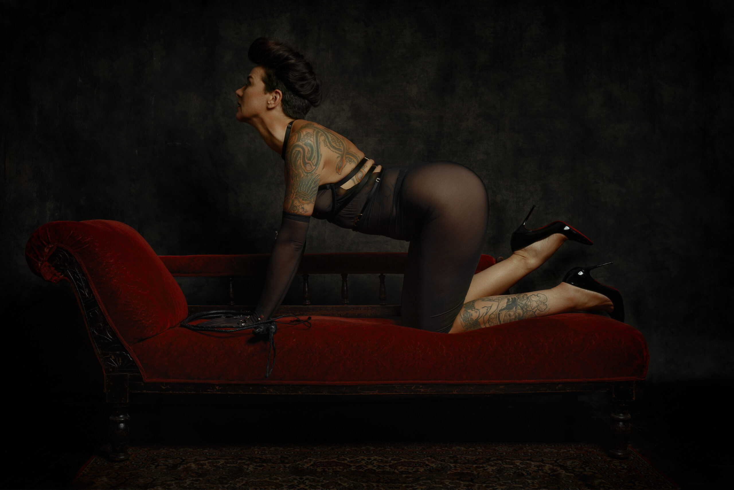 Fine art photograph of Miss V kneeling on a red velvet chaise longue, tattooed, in a sheer black dress and heels, dramatic low-key lighting — by Menno van der Meulen.