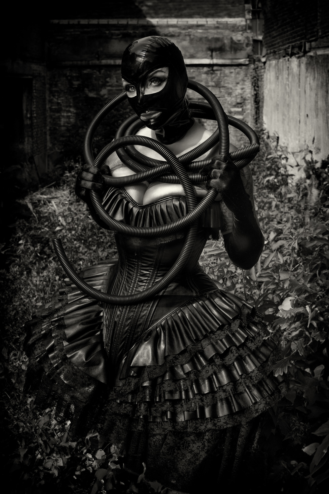 Apocalypse Garden #2 Black-and-white fine-art portrait of a masked woman entwined with industrial hoses in an overgrown yard — photo by Menno van der Meulen.