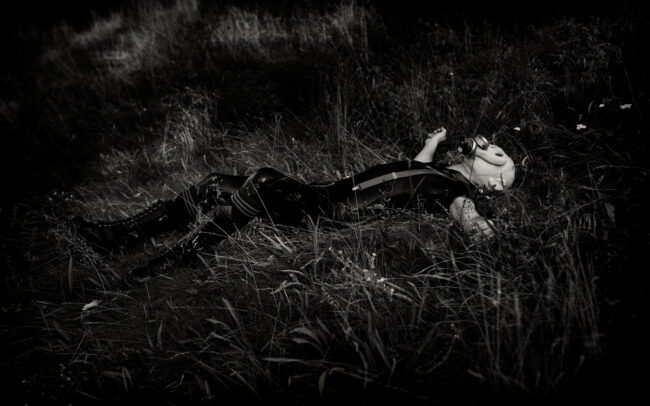 Latex-clad body wearing a gas mask and lying in a field, dark fetish survival atmosphere, photographed by Menno van der Meulen
