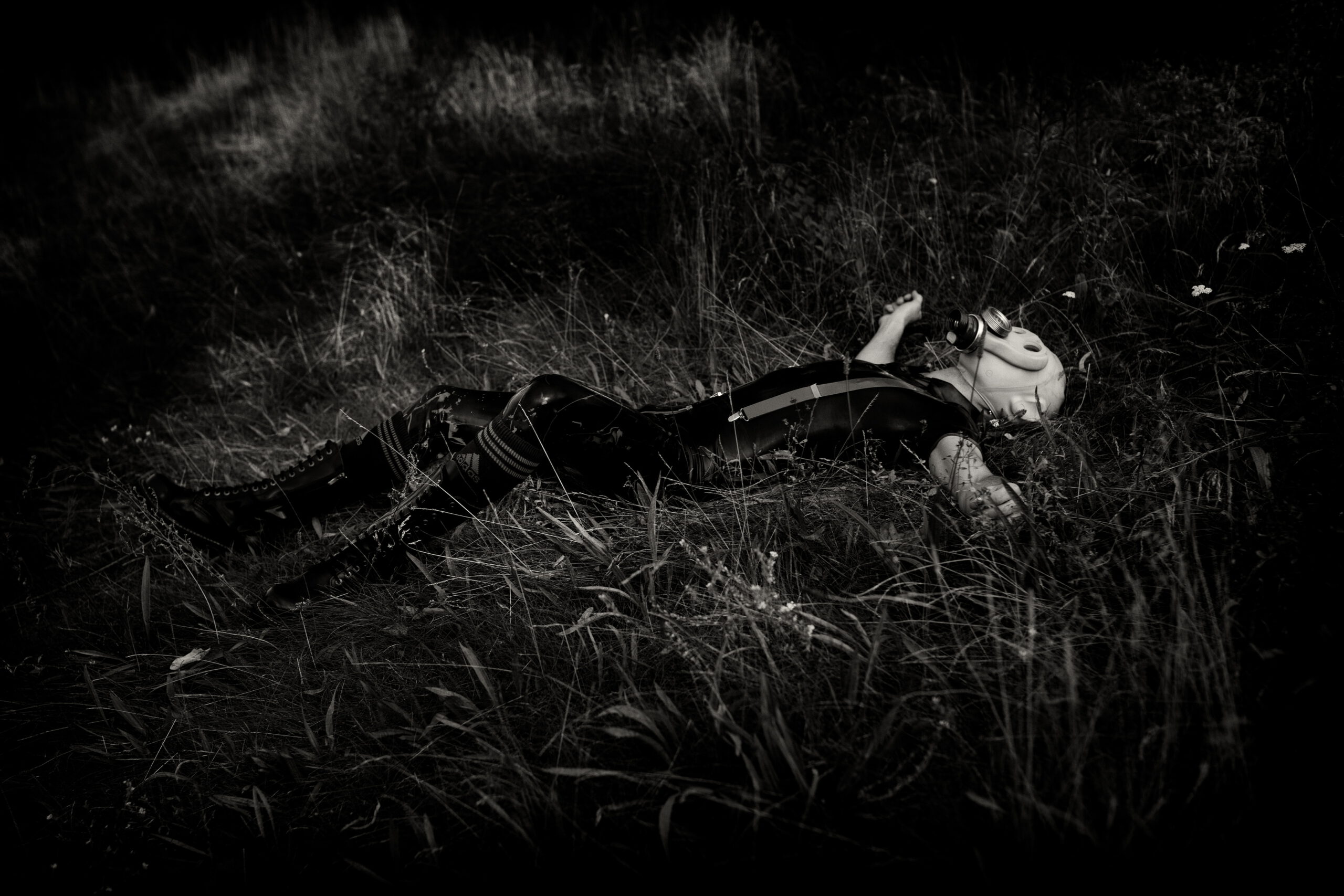 Latex-clad body wearing a gas mask and lying in a field, dark fetish survival atmosphere, photographed by Menno van der Meulen