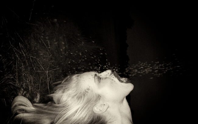Fine art black-and-white photograph of a woman lying by tall grass, head tilted back and exhaling a spray of droplets into the night — by Menno van der Meulen
