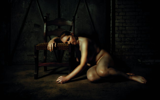 Nude female figure resting on the floor beside a vintage chair in a dark industrial room, cinematic light on the skin. Fine art photograph by Menno van der Meulen.