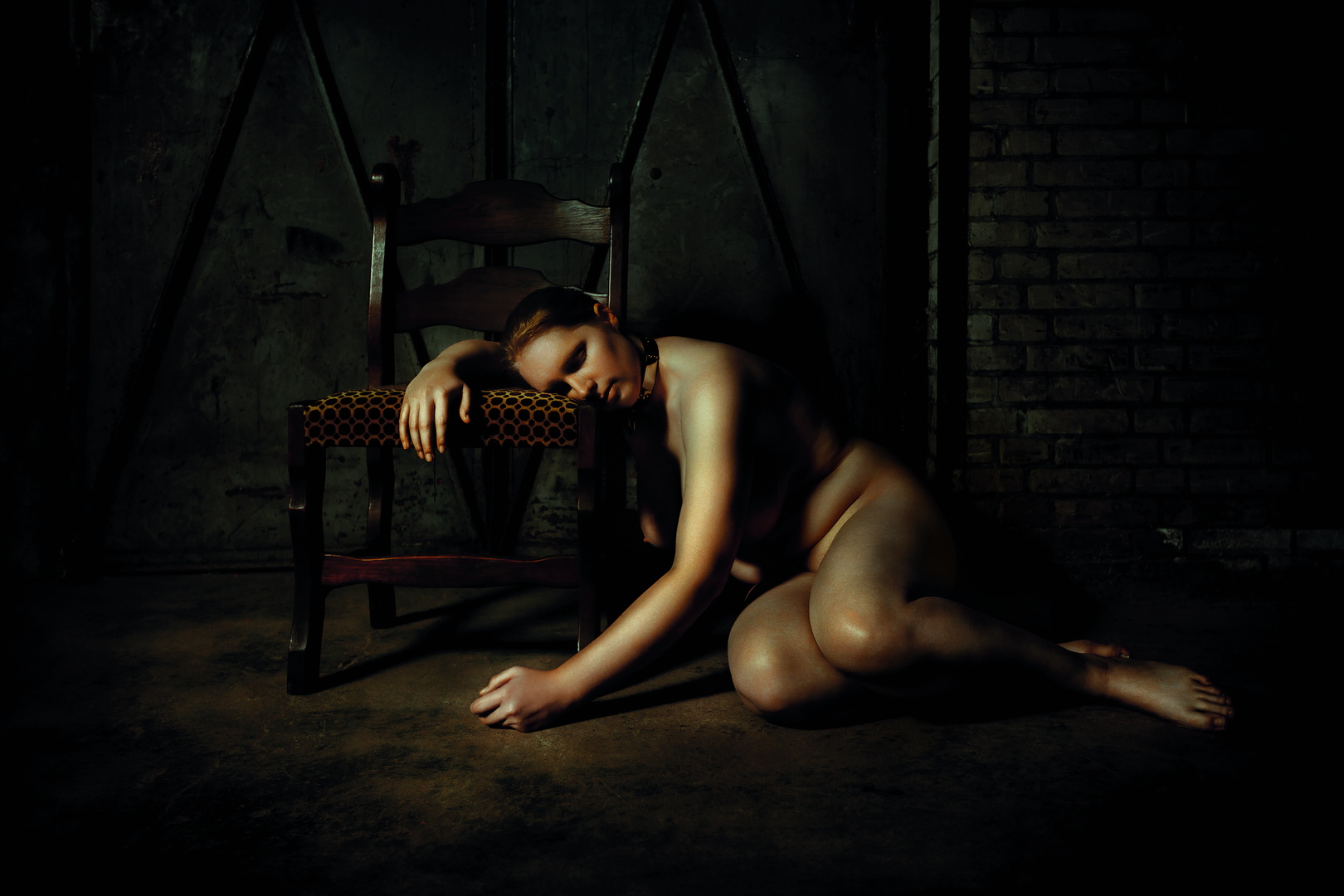 Nude female figure resting on the floor beside a vintage chair in a dark industrial room, cinematic light on the skin. Fine art photograph by Menno van der Meulen.