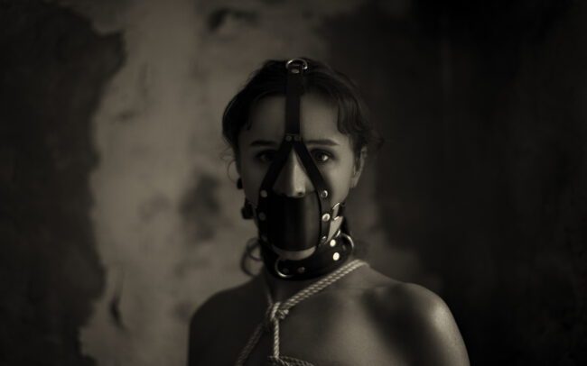 Fine art black-and-white portrait of a woman wearing a leather muzzle mask and collar, intense eyes and moody light — by Menno van der Meulen.