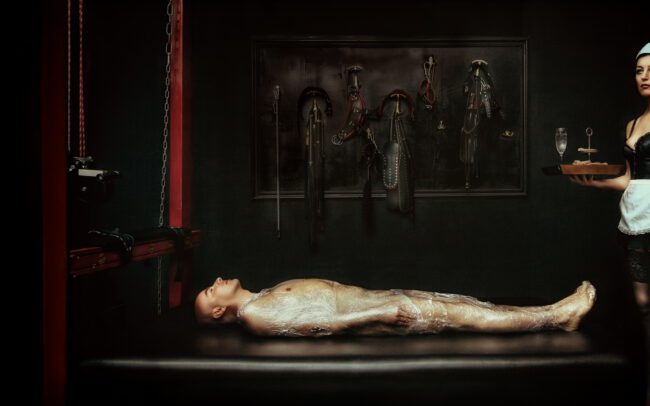 A restrained nude male figure wrapped in plastic on a clinical bondage table while a uniformed attendant holds a serving tray of restraints; an image about control, ritual and care by Menno van der Meulen.