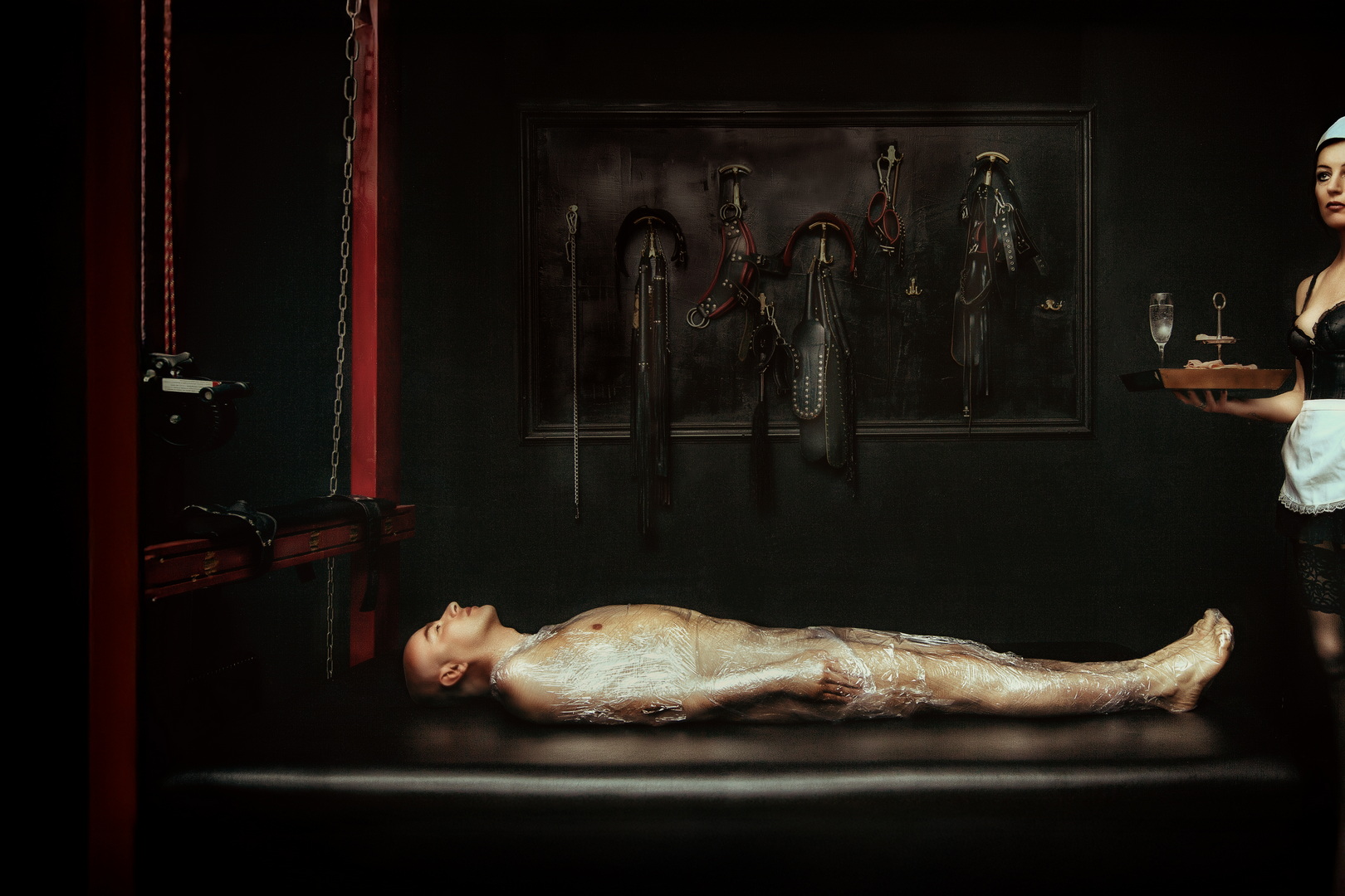 A restrained nude male figure wrapped in plastic on a clinical bondage table while a uniformed attendant holds a serving tray of restraints; an image about control, ritual and care by Menno van der Meulen.