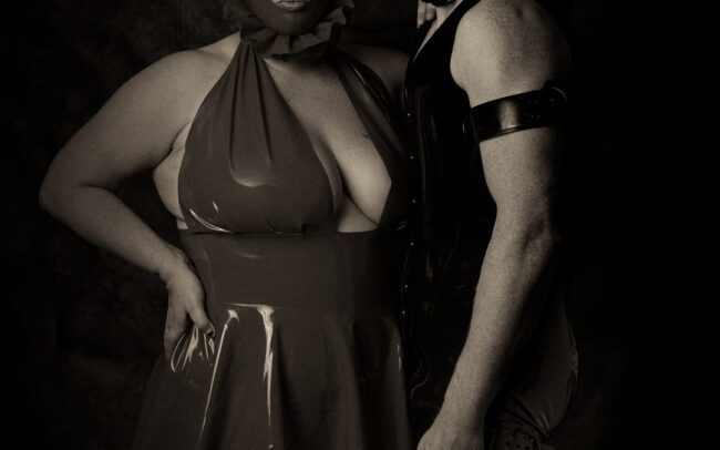 Two masked figures standing together in a studio, wearing dark clothing and looking toward the camera.